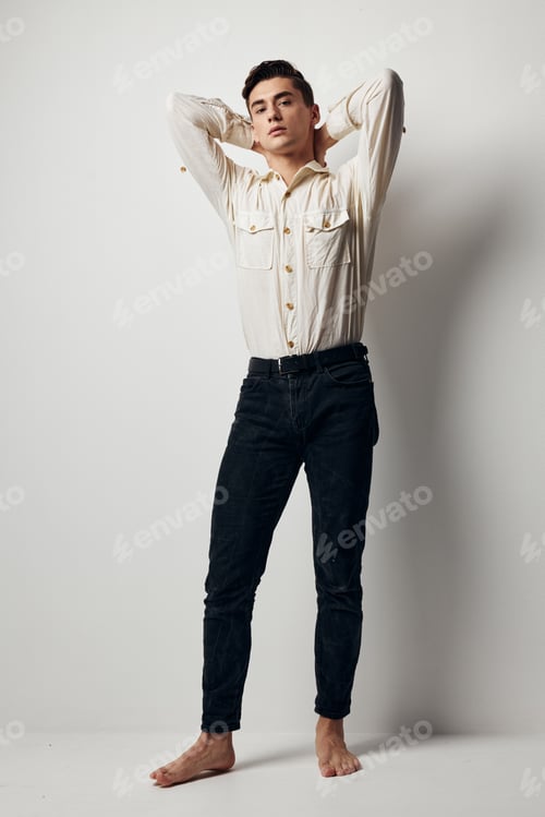 Preview: A handsome guy in a white shirt and trousers holds his hands behind his head on a light background