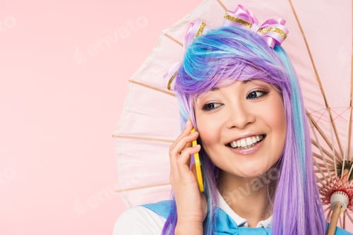 Preview: Smiling asian anime girl holding paper umbrella and talking on smartphone isolated on pink