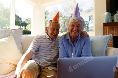 Preview: Senior couple wearing party hats leaning toward laptop on couch in living room with lit candle