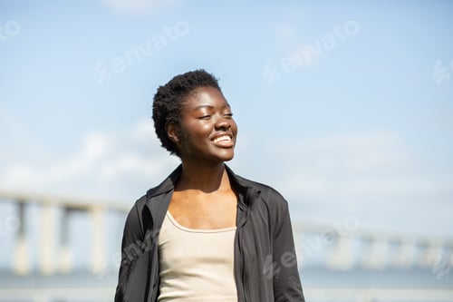 Preview: Smiling young woman enjoying a sunny day near a waterfront bridge