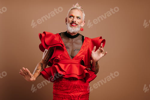 Preview: Mature gay man with crown on head wearing beautiful red dress and dancing against brown background