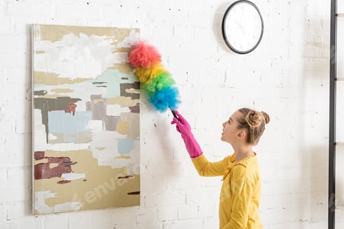 Preview: Cute child in rubber gloves sweeping away dust from picture with feather duster in living room