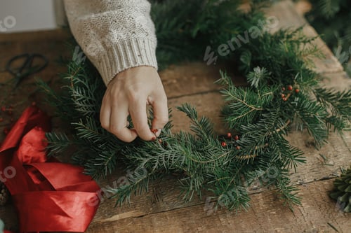 Preview: Making Christmas wreath. Woman hands holding red berries and decorating wreath on wooden table