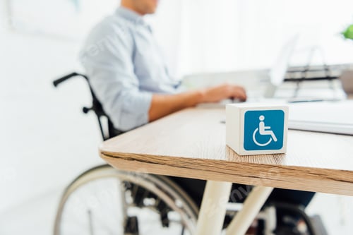 Preview: Selective focus of cube with disabled sign and man in wheelchair sitting at table