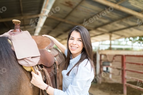 Preview: Portrait Of Charming And Smiling Female Rider Preparing Horse Saddle At Stable