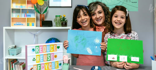 Preview: Teacher in classroom looking at camera next to students showing colorful ecological drawings