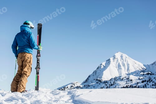 Preview: Skier standing in snow, holding skis, looking at mountain, rear view