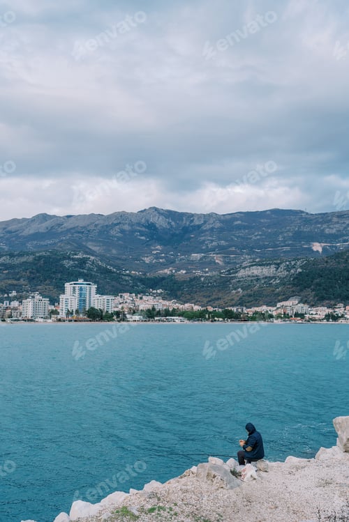 Preview: Man with a fishing rod sits on a rocky slope above the sea and catches fish. Side view