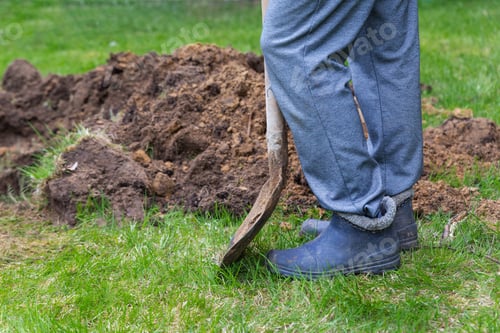 Preview: Legs of a man in rubber boots with a shovel near the excavated ground close-up.