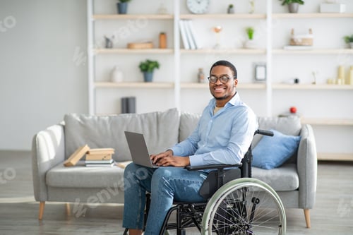Preview: Happy disabled black man in wheelchair using laptop, working online from home, copy space