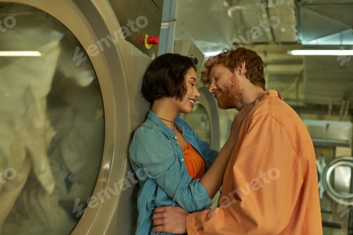 Preview: smiling multiethnic romantic couple kissing near washing machine in public laundry