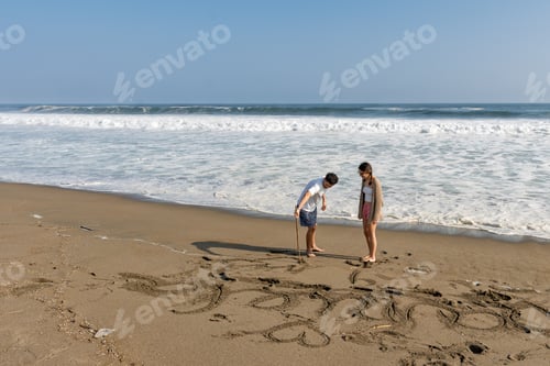 Preview: Young couple drawing name hearts on beach sand