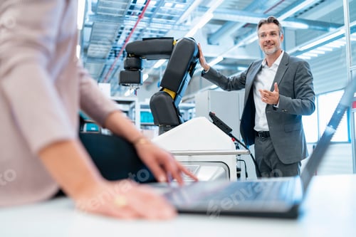 Preview: Businessman and businesswoman with laptop and robot in a modern factory hall