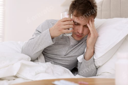 Preview: Unhappy young man with glass of water suffering from hangover on bed at home