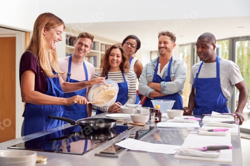 Preview: Female Teacher Making Pancake On Cooker In Cookery Class As Adult Students Look On