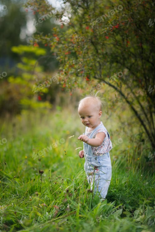 Preview: funny emotional toddler baby in the summer in the park looks at flowers and blades of grass