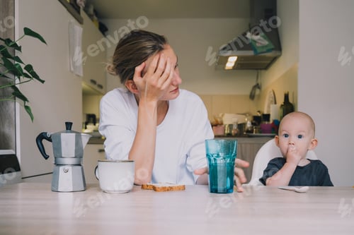 Visualização: Jovem exausta com bebê está sentada com café na cozinha.