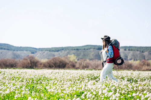 Preview: Woman with red backpack and hat walking through flowery field