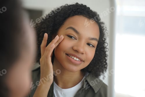 Preview: Biracial young girl looks at mirror, applying moisturizing facial cream. Natural beauty, skincare