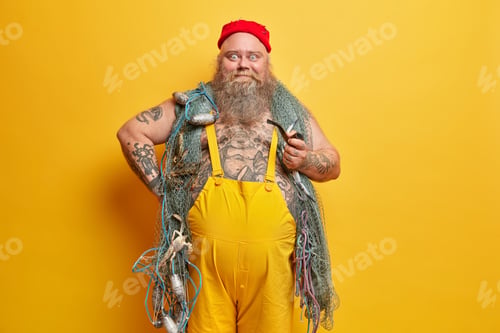 Preview: Positive bearded fisherman ready for fishing, holds smoking pipe, wears red hat and overalls, enjoys
