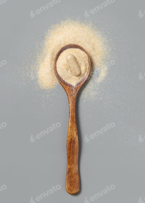 Preview: Wooden spoon full of beige powder and capsule on grey top view. Dietary supplement or kitchen spice
