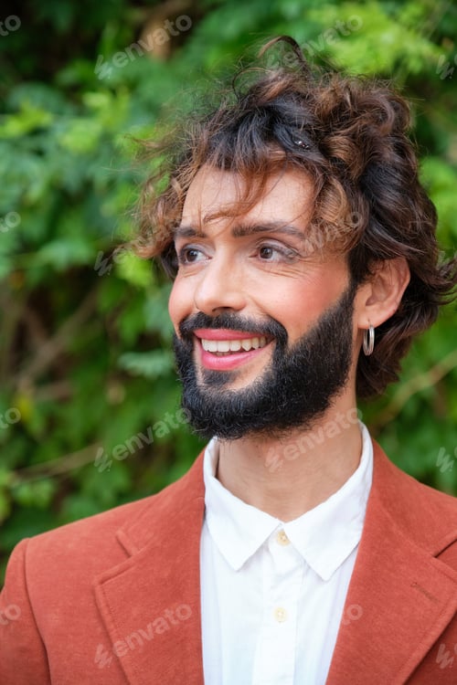 Preview: Man with makeup looking away and smiling while standing outdoors against green leaves background.