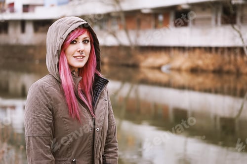 Preview: pierced young woman with pink hair and hooded parka standing by river
