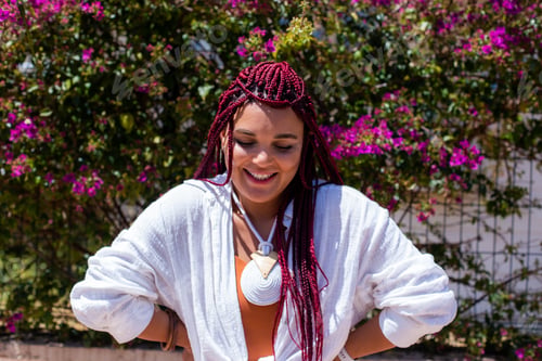 Preview: Diverse beautiful woman with dreadlock in white shirt smiling and spending time by purple bush