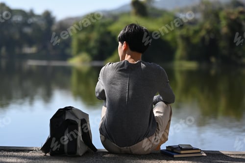 Preview: Back view of man sitting alone near lake with mountains sunny landscape on background.