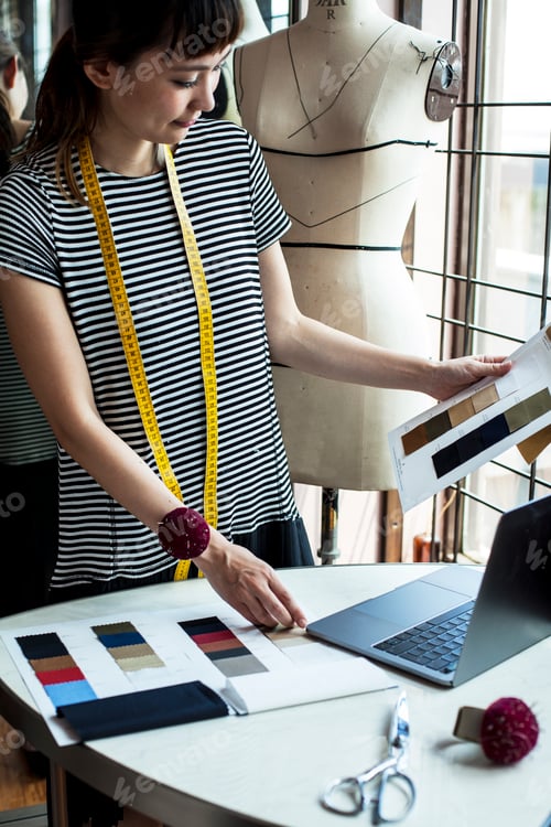 Preview: Japanese female fashion designer working in her studio, looking at fabric samples, using laptop.