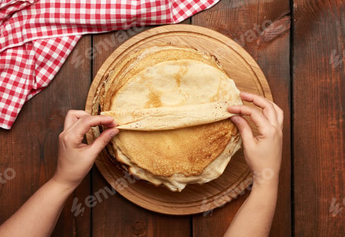 Preview: Fried round pancakes on a wooden board and two female hands, brown table