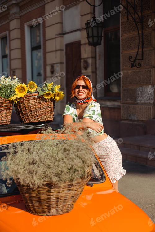 Preview: A young woman on a summer day stands leaning on an orange vintage car