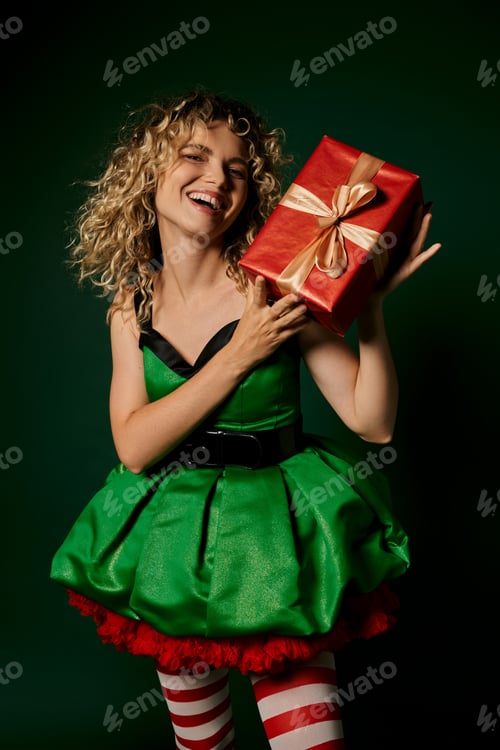 Preview: cheerful woman in festive dress holding present in hands and smiling at camera, new year elf concept