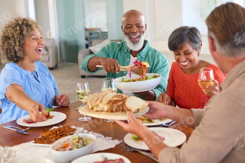 Preview: Group Of Smiling Mature Friends At Home Sitting Around Table Meeting For Lunch And Wine Together