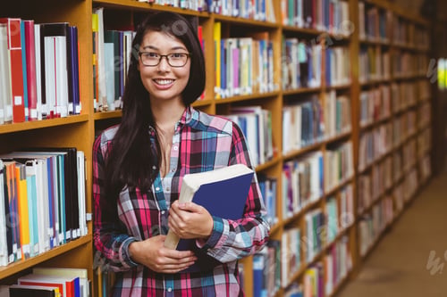 Preview: Asian woman in plaid shirt leaning against bookshelf holding blue book in library aisle, copy space