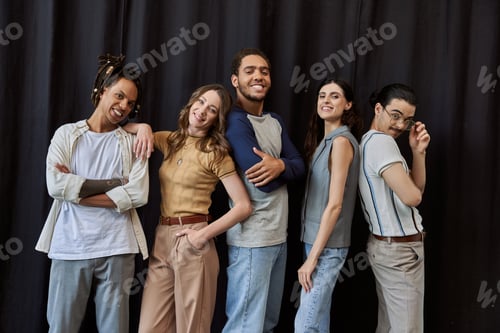 Preview: joyful multiethnic colleagues in casual attire looking at camera near black drape in modern office