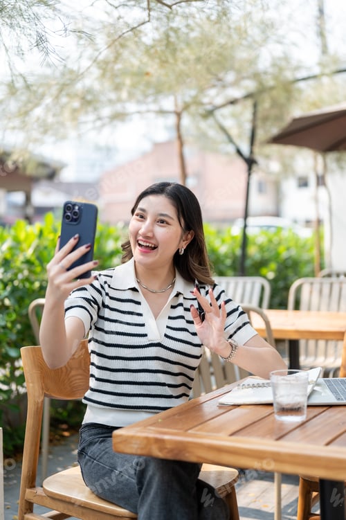 Preview: Pretty asian woman waving a hand to video call on phone while sitting at wooden table in garden cafe