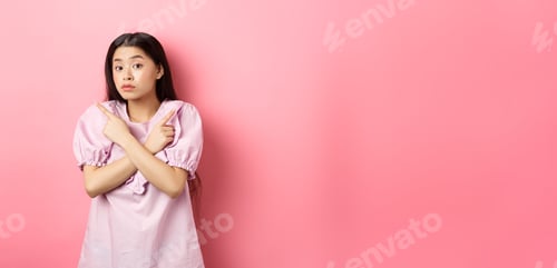 Preview: Clueless cute asian girl pointing sideways and look confused, cant make decision, standing hesitant