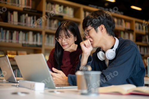 Preview: Glasses asian man student and a woman friend looking at phone together while sits at library's table