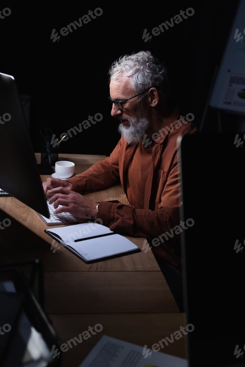 Preview: bearded grey haired businessman typing on computer near notebook at night in office