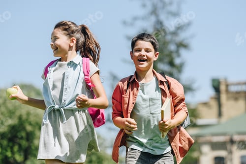 Preview: two happy schoolkids with backpacks smiling while running in park