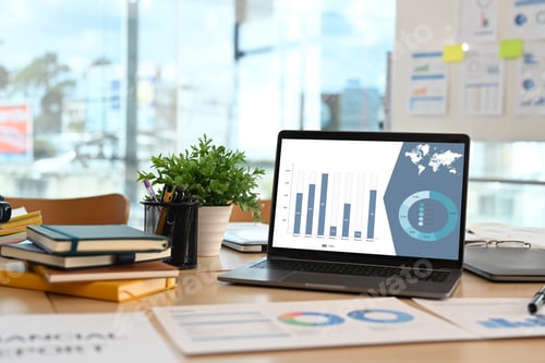Preview: Front view of laptop with financial graph data on screen and office supplies on wooden office desk.