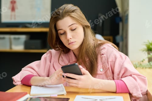 Preview: Teenage female student in pink jacket looking at smartphone screen at lesson