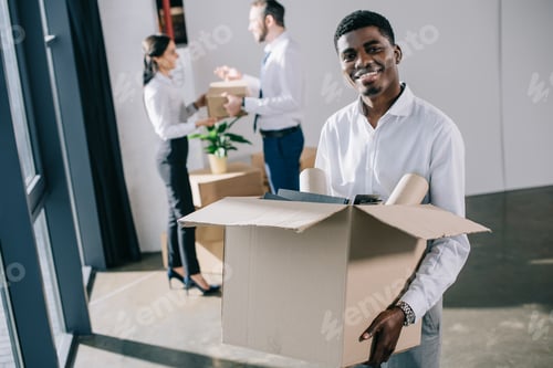 Preview: african american businessman holding cardboard box and smiling at camera while colleagues standing