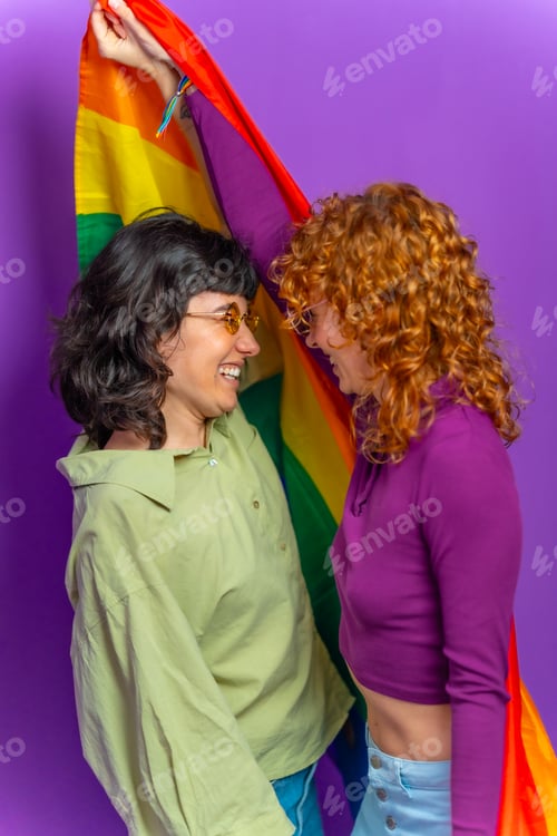 Preview: Lesbian couple about to kiss, smiling raising lgbt rainbow flag