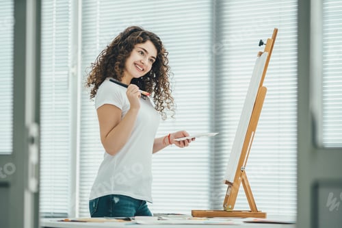 Preview: The happy curly woman painting a picture on the easel.