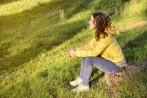 Preview: young brunette girl smiling with long curly hair in profile sitting on a tree trunk