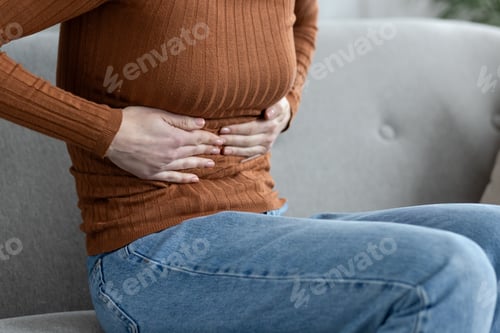 Preview: Young female hands holding her stomach, woman sitting on sofa at home feeling severe pain and