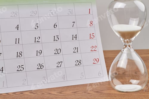 Preview: Close-up hourglass and month calendar on wooden desk.