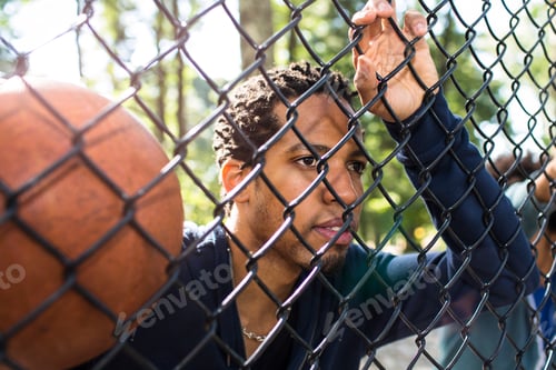 Preview: Portrait of young man holding basketball through wire fence